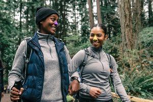 Two smiling black women link arms as they trek through the forest. They are wearing long-sleeved winter clothes and colorful sunscreen for sun protection.